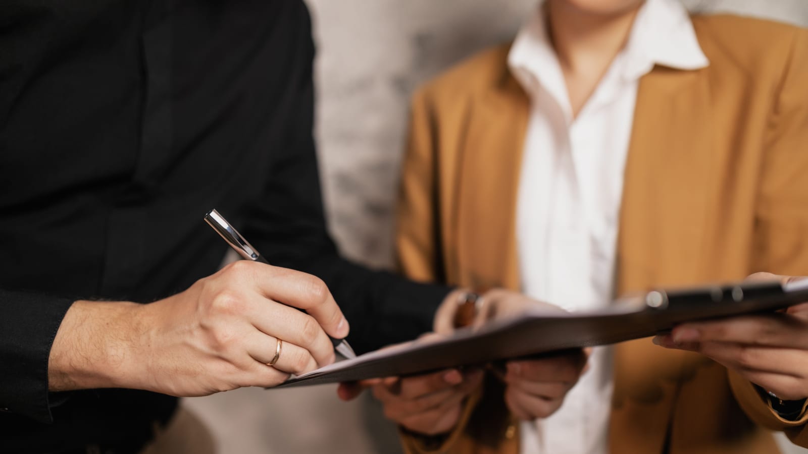 One person signs a page on a clipboard held by another, like a written franchise approval. A silver pen is in hand, and both faces are out of frame.