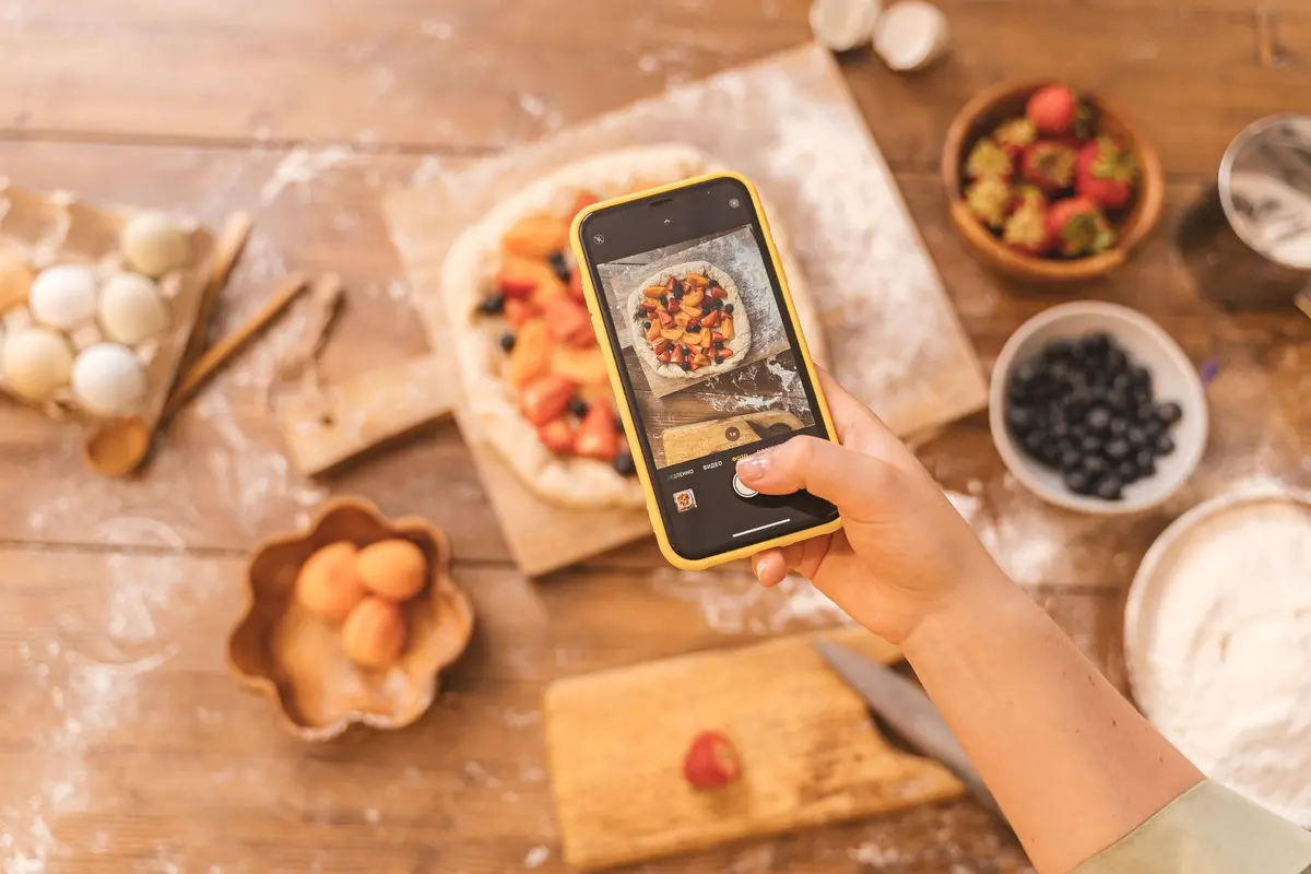 A hand holds a phone in a yellow case over a fruit tart on a floured wooden board, framing the shot on screen beside bowls of berries and apricots.