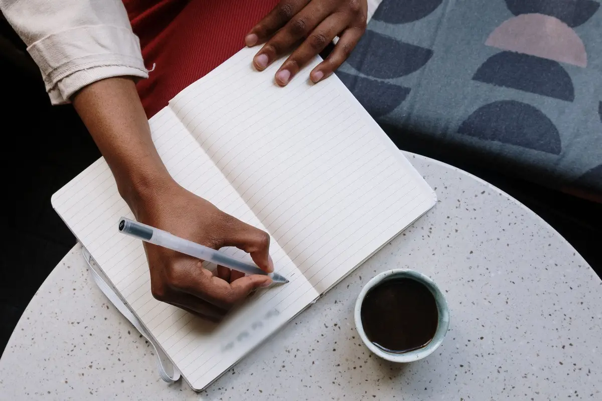 Seen from above, a hand writes with a clear pen in an open lined notebook on a speckled round café table, next to a cup of black coffee.
