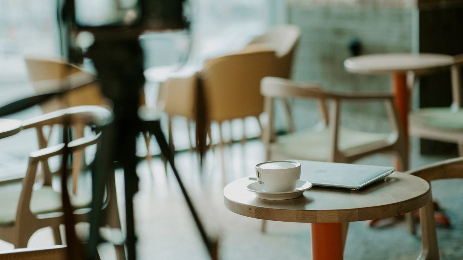 A video camera on a tripod stands ready to film in a quiet cafe dining room, aimed past an empty table with one coffee cup.