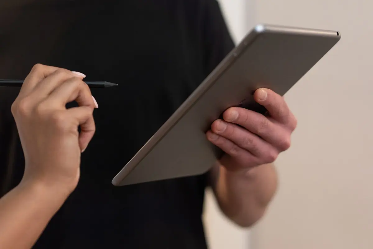 One person holds out a tablet while another holds a stylus, ready to sign. Faces are out of frame.