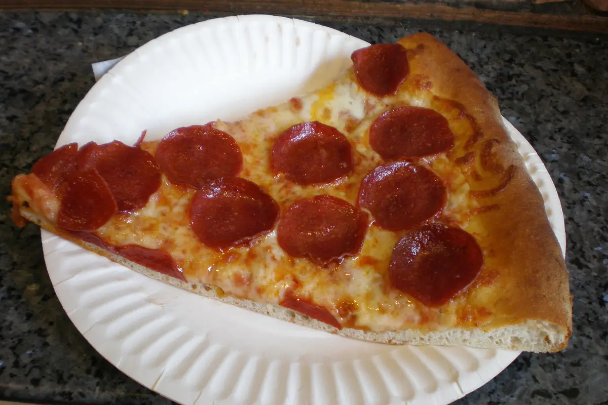 A pepperoni pizza slice on a white paper plate, the way a concession stand sells it.