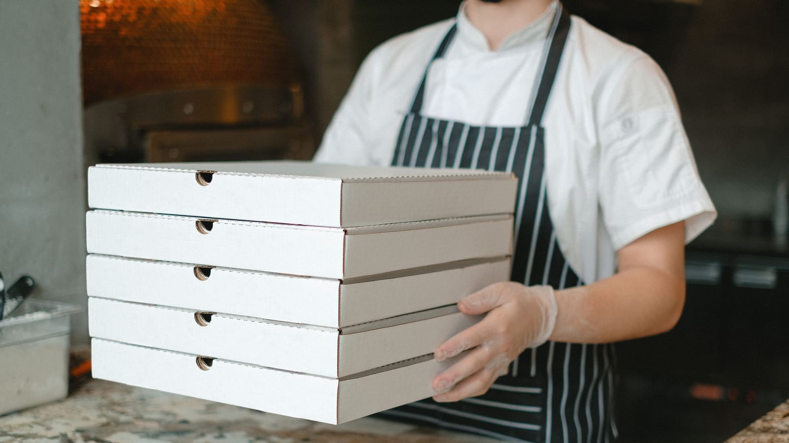 A pizza shop worker holds a stack of plain pizza boxes, the kind of pickup a high-school team dinner needs.