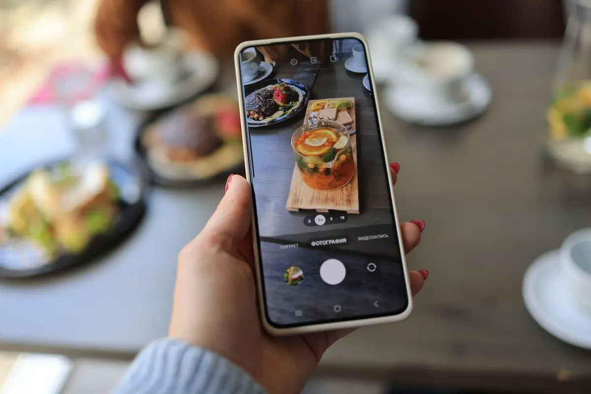 A hand holds a phone over a cafe table to photograph a plate of food and two drinks.