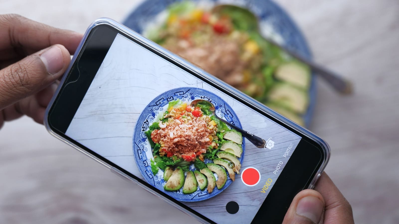 Two hands hold a phone in video mode over a plate of salad with avocado slices, filming the food the way food influencers do.