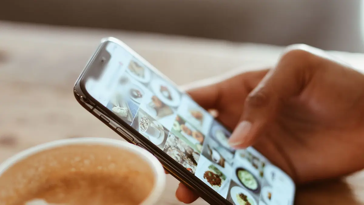 A hand scrolls a phone through a grid of food pictures, beside a cup of latte.