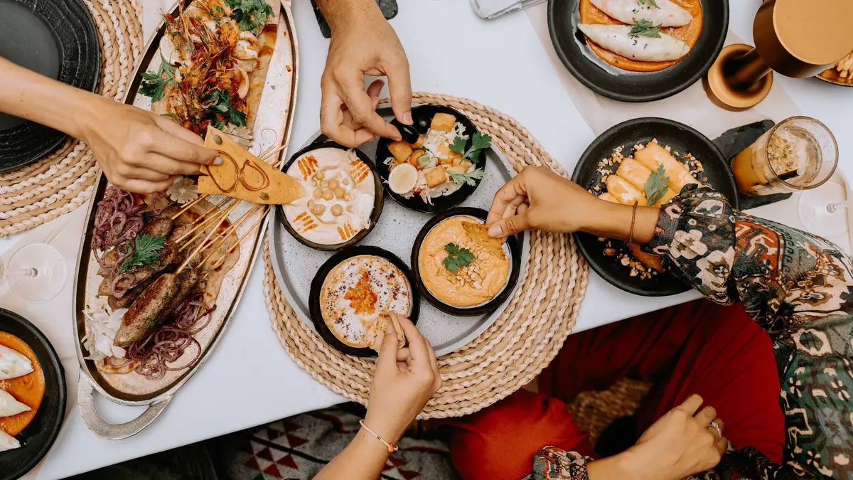 Friends’ hands reach across a table of shared plates, dipping chips into hummus beside skewers and dumplings.