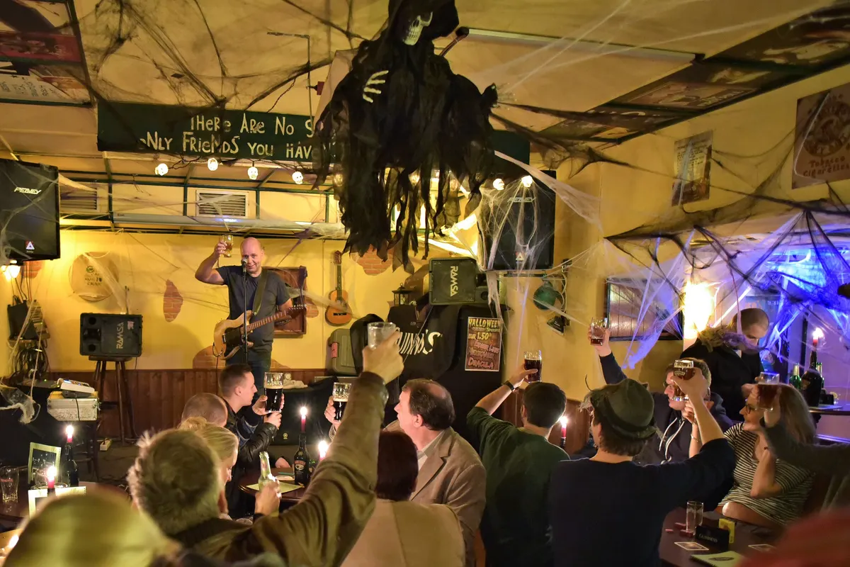 Guests raise their glasses in a pub hung with cobwebs and a grim reaper, while a guitarist plays.