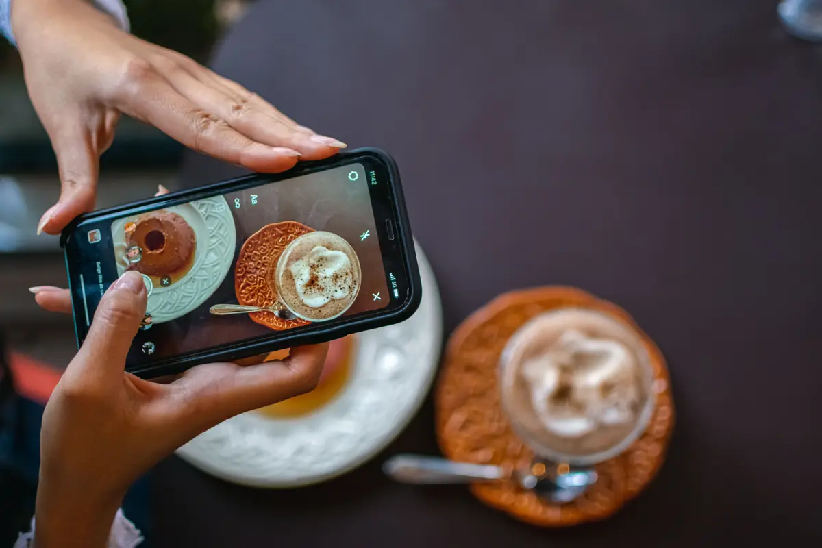 Two hands hold a phone above a cafe table and frame a dessert and a frothy drink on its screen.