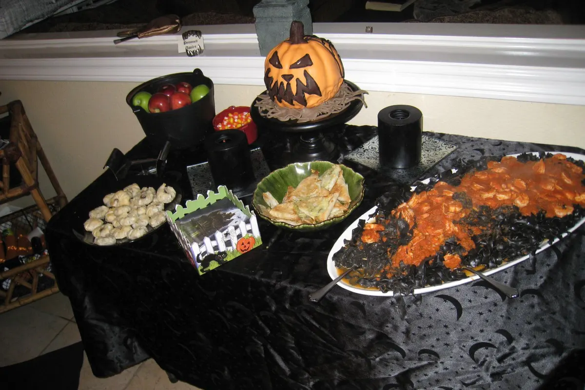 A Halloween party table at home, with black pasta and shrimp, dumplings, candy corn and a pumpkin cake.