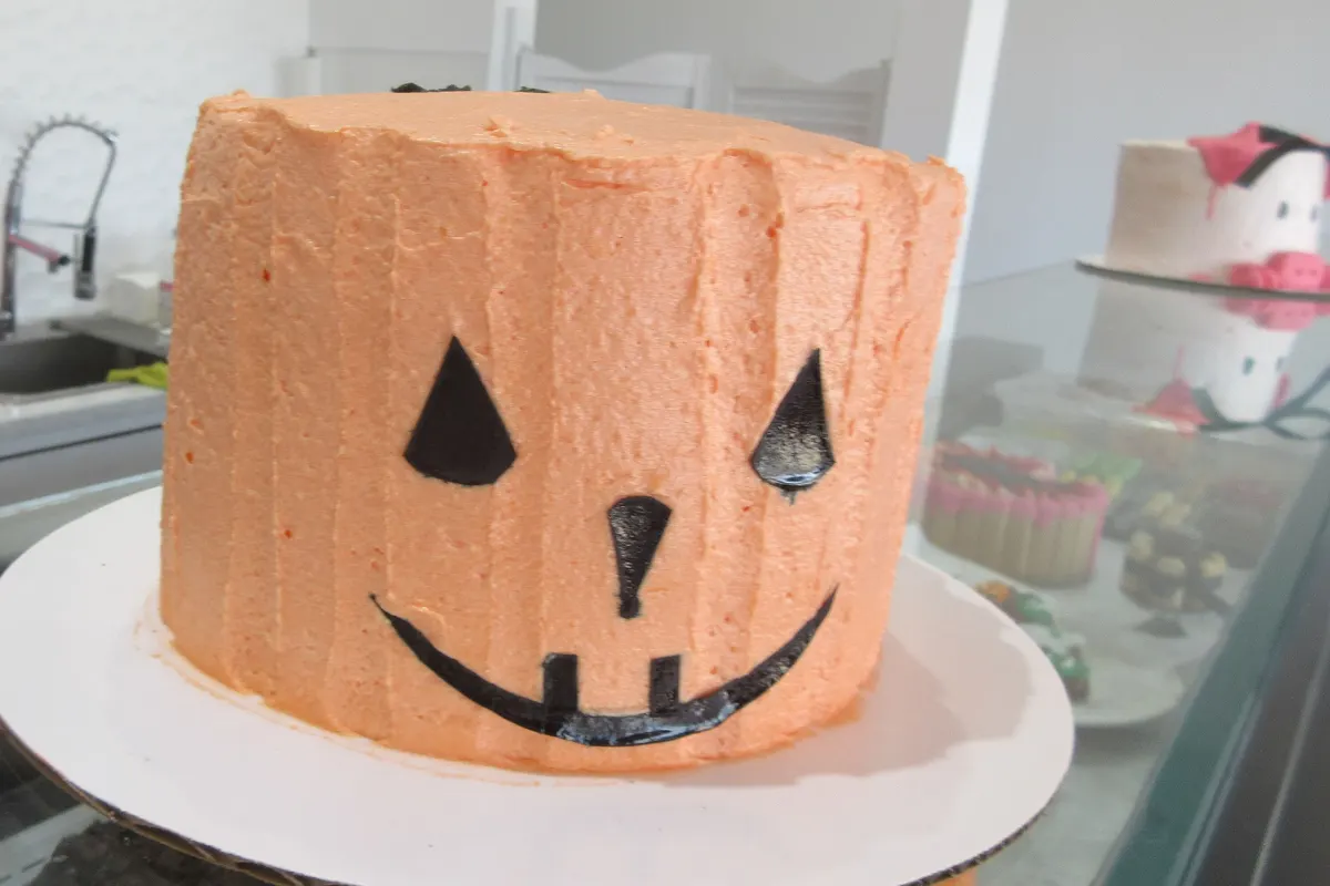 A tall cake frosted orange with a black jack-o’-lantern face, on a bakery counter.
