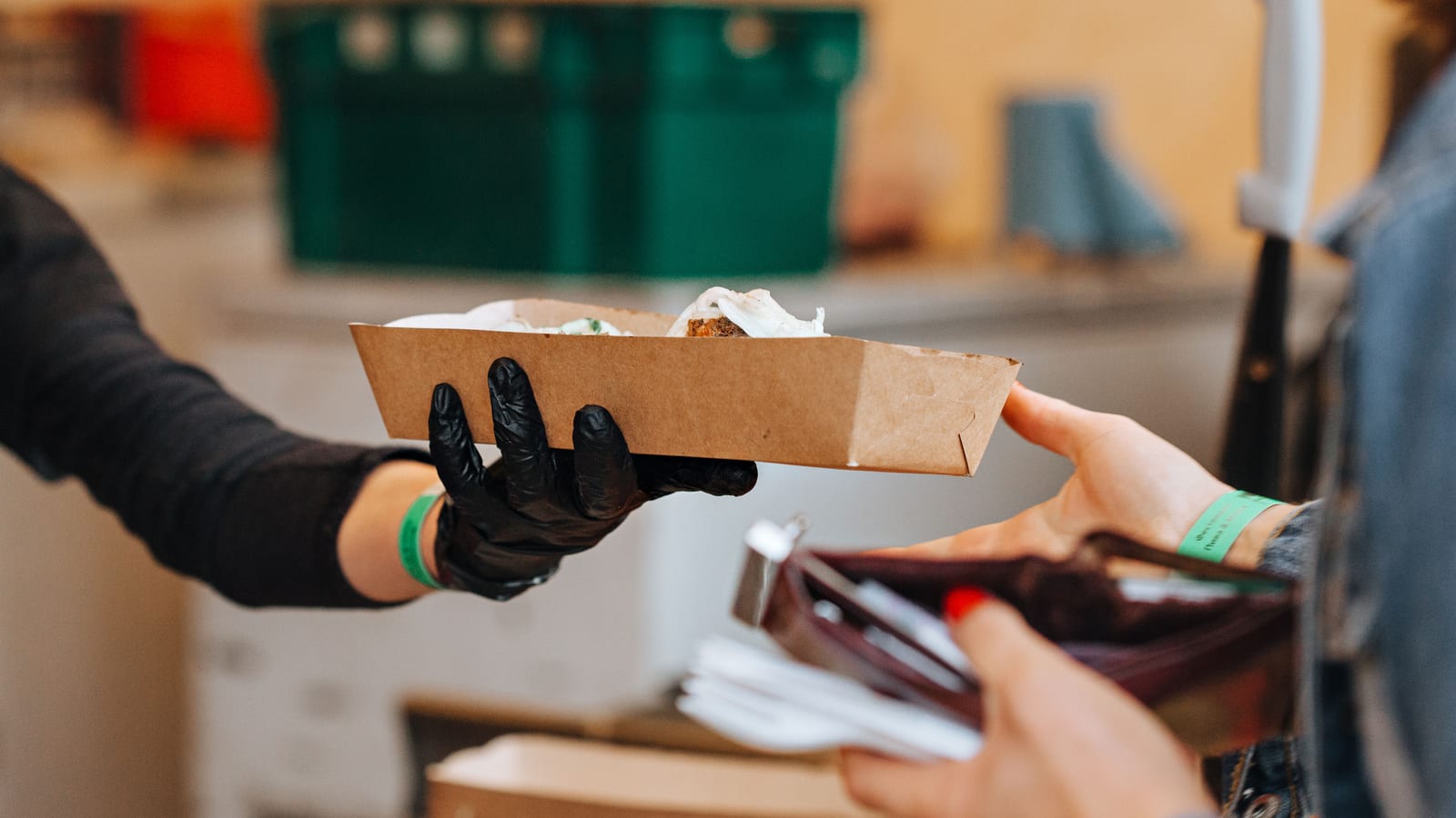 A gloved hand passes a takeout tray of food across a counter to a guest holding an open wallet.