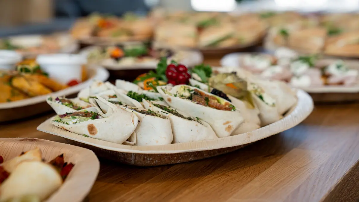 A round platter of rolled wraps with greens and red currants sits on a wooden table among other catering trays.