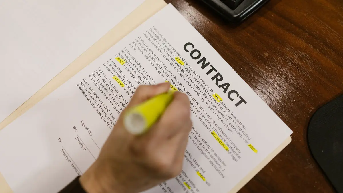 A hand marks lines on a printed contract with a yellow highlighter, on a wooden desk.