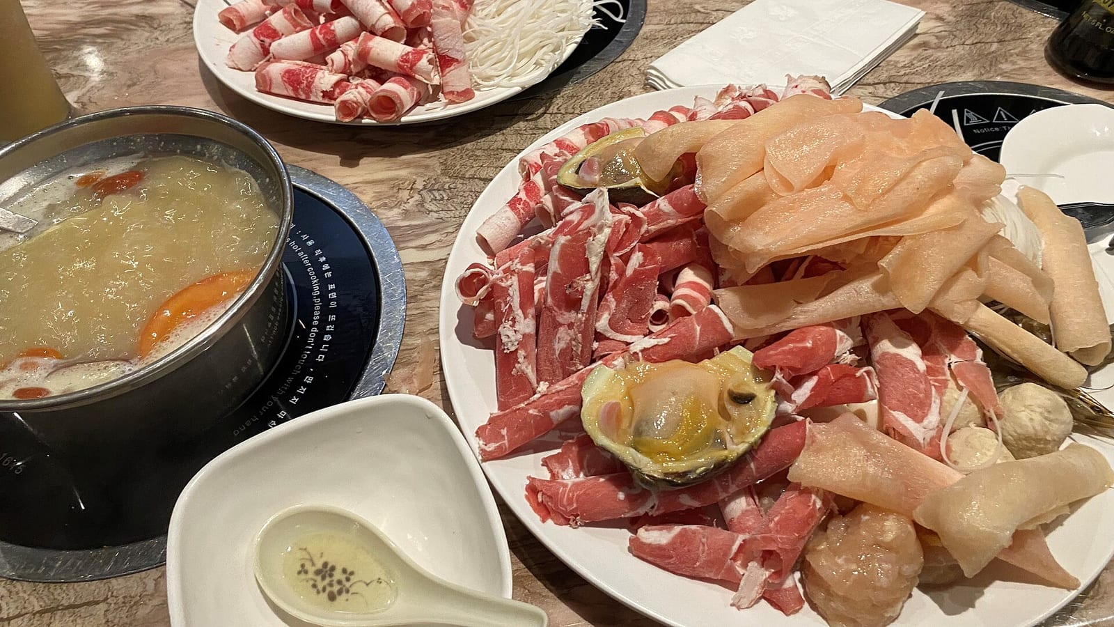 A hot pot table set for dinner, with broth on a burner, platters of rolled thin-sliced meat and seafood, and small sauce dishes.