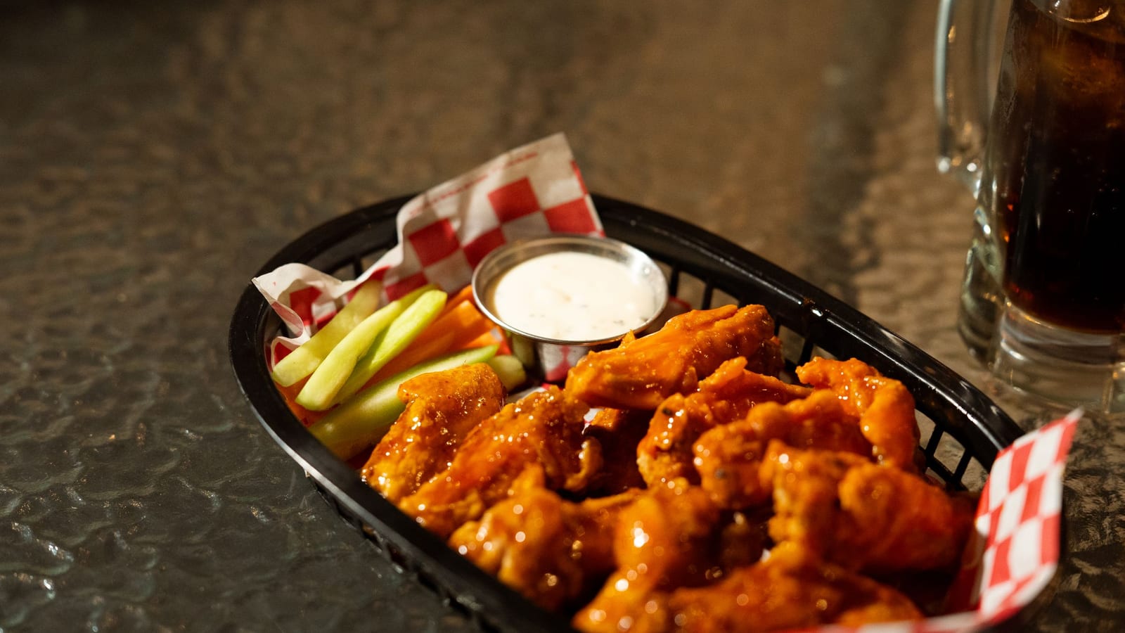 A basket of glazed chicken wings with celery, carrot sticks and a cup of ranch sits on checkered paper on a bar table, next to a mug of soda.