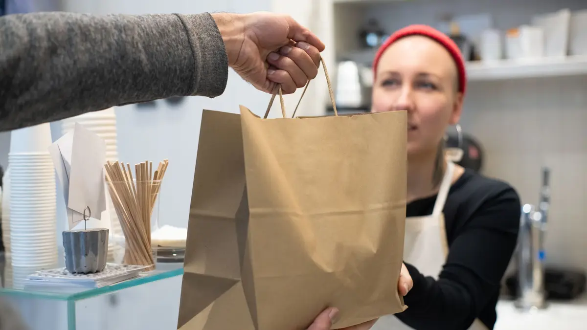 A worker in an apron hands a paper takeout bag across the counter to a customer.