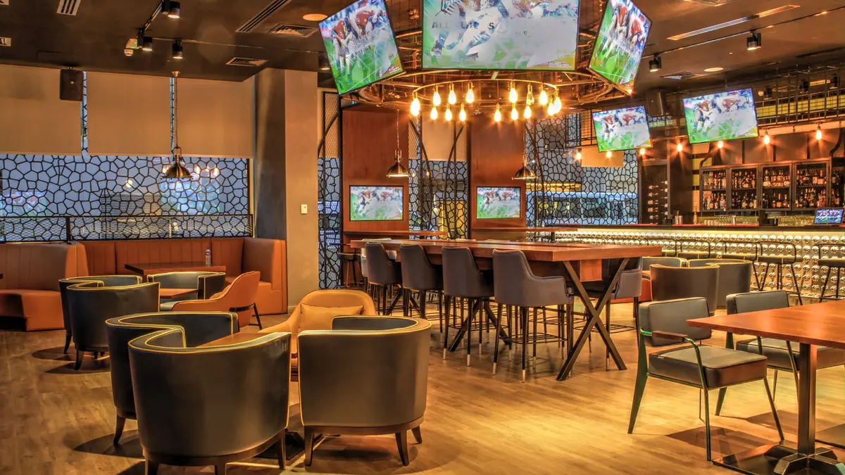 An empty sports bar dining room, set before opening, with lounge chairs, high tables and a football game on many screens.