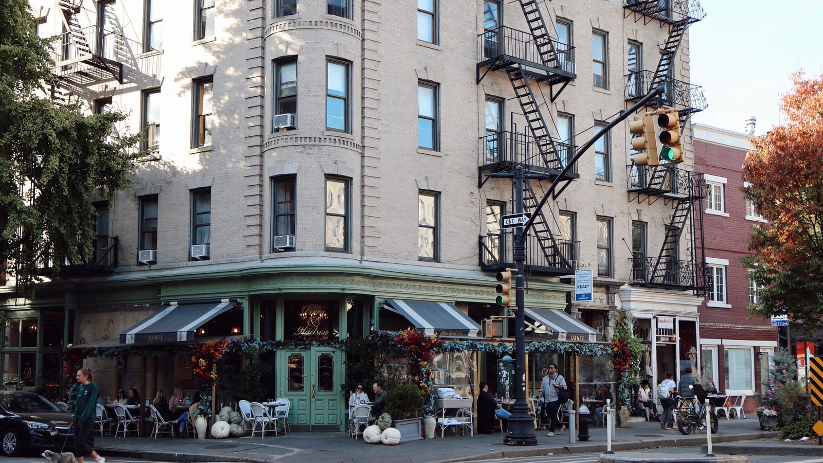 A corner restaurant in a New York City walk-up building, with fire escapes above and diners at sidewalk tables on an autumn afternoon.