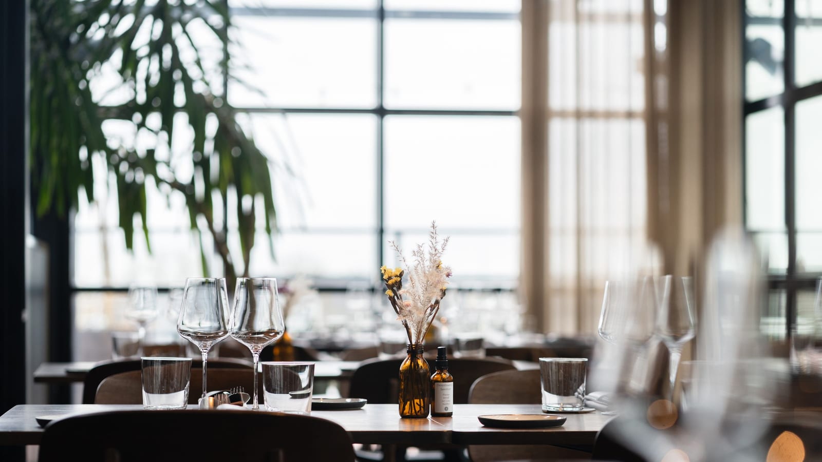 A restaurant dining room before service: tables set with wine glasses and plates, dried flowers in a vase, tall windows behind.