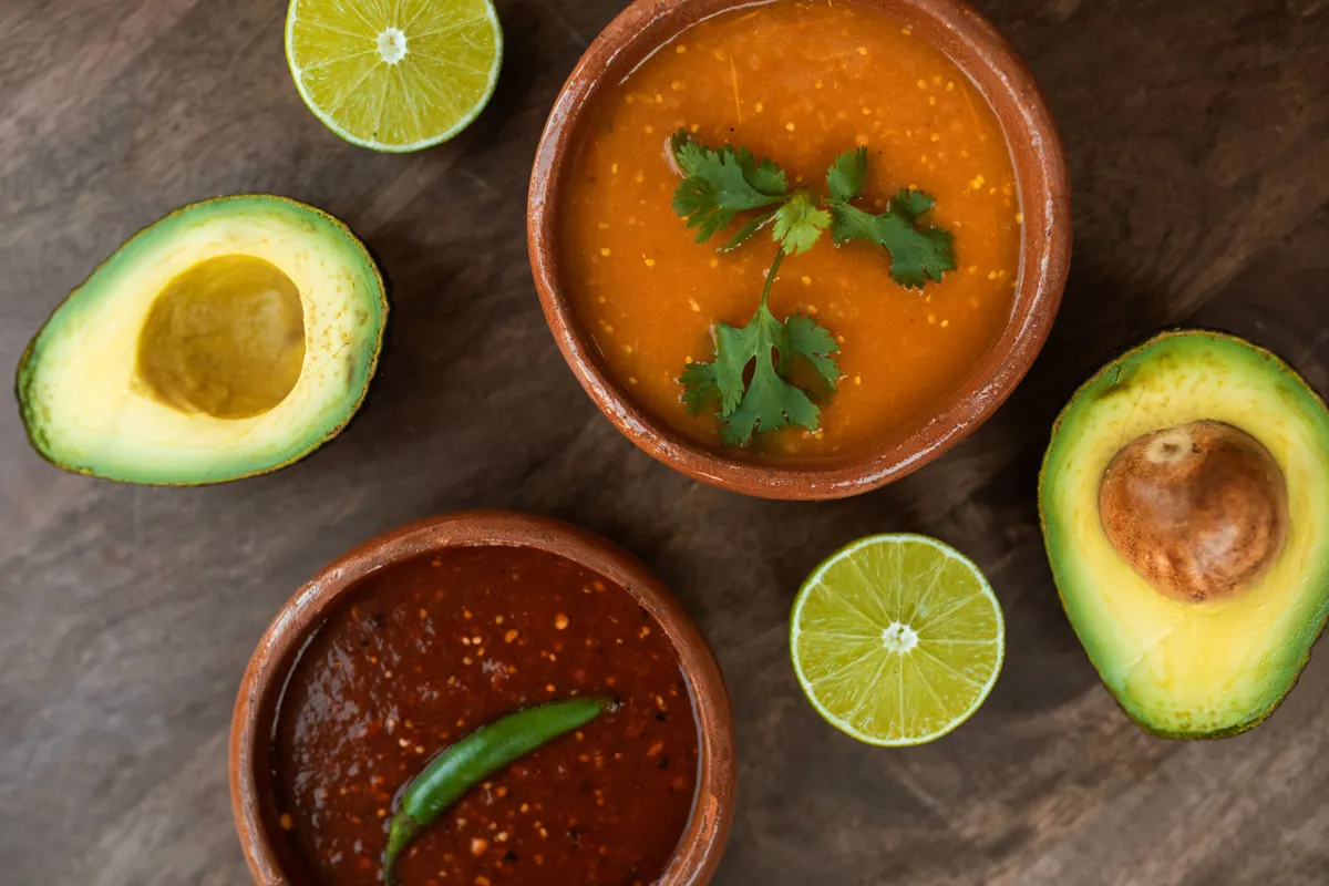 Two salsas, one orange with cilantro and one dark red with a green chile, sit in clay bowls beside halved limes and avocados.
