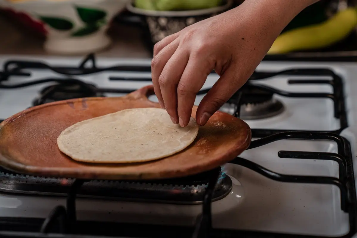 A hand lays a raw corn tortilla on a clay comal over a gas burner.