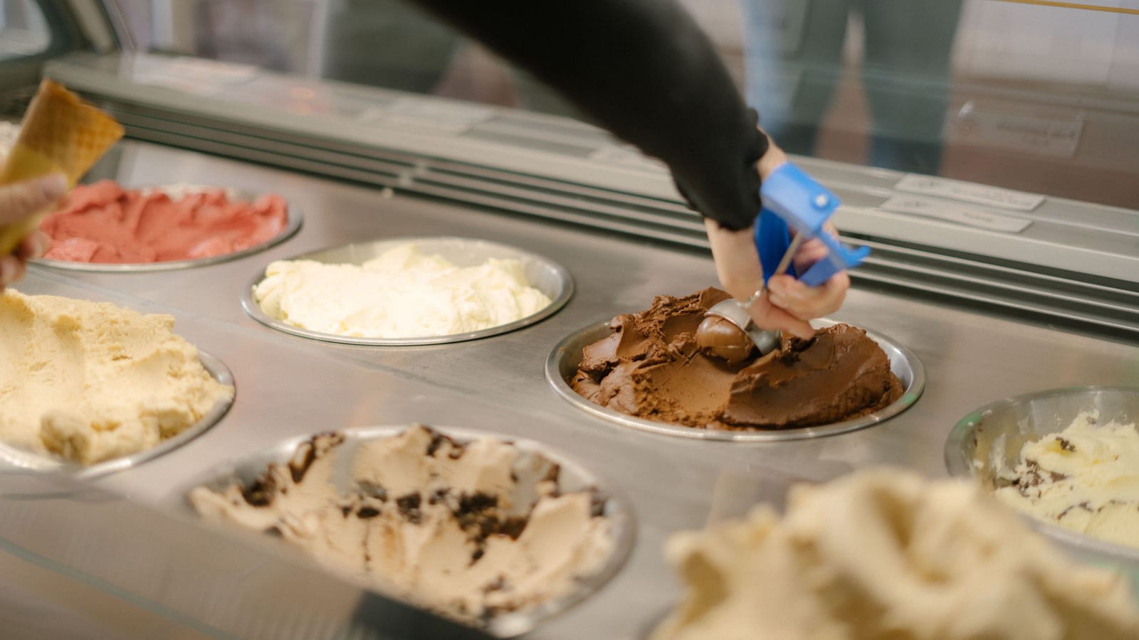A worker in a long black sleeve scoops chocolate ice cream from a round tub in a dipping cabinet, while another hand holds out a waffle cone.