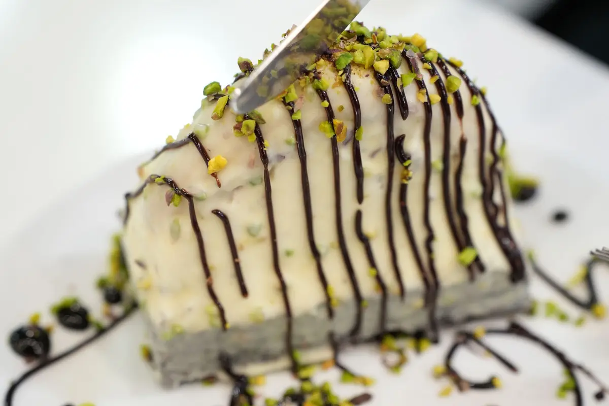 A knife cuts into a slice of pistachio ice cream cake striped with chocolate sauce and chopped pistachios.