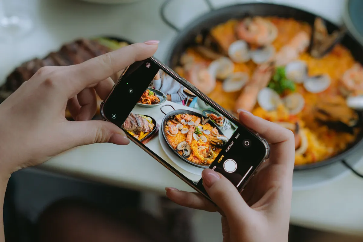 Hands hold a phone over a restaurant table and shoot a pan of seafood paella.
