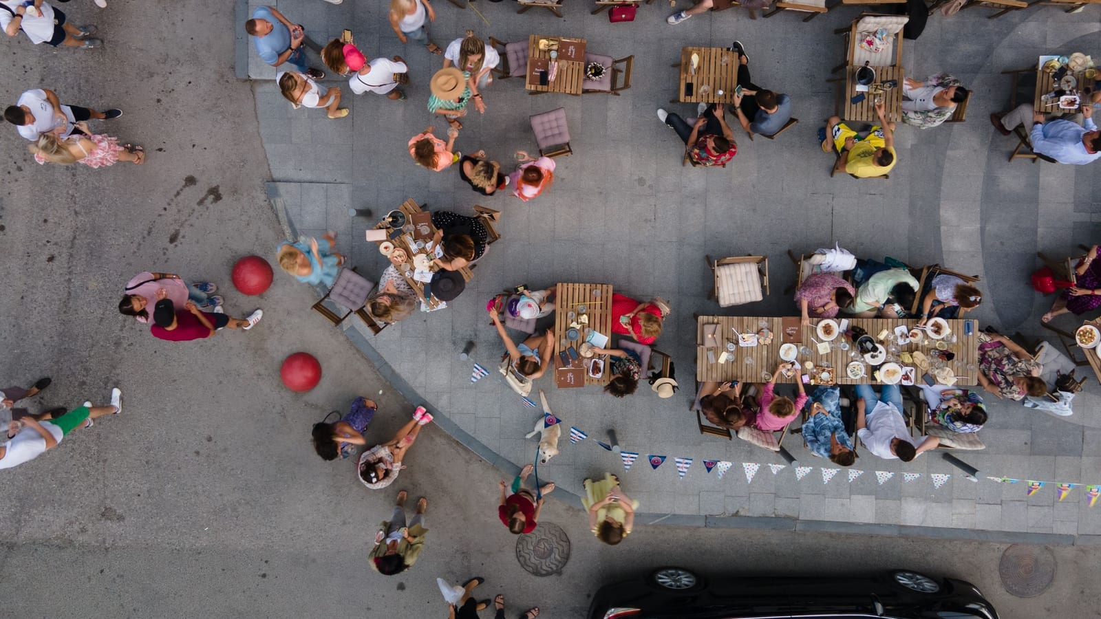 An overhead view of a sidewalk café on a city street, where neighbors share meals at wooden tables while people walk past with a small dog under a line of bunting.