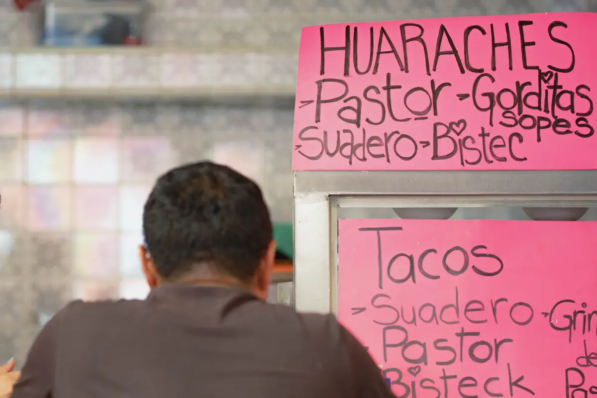 Hand-lettered pink signs over a taquería counter list huaraches, gorditas, sopes and tacos de pastor, suadero and bistec.
