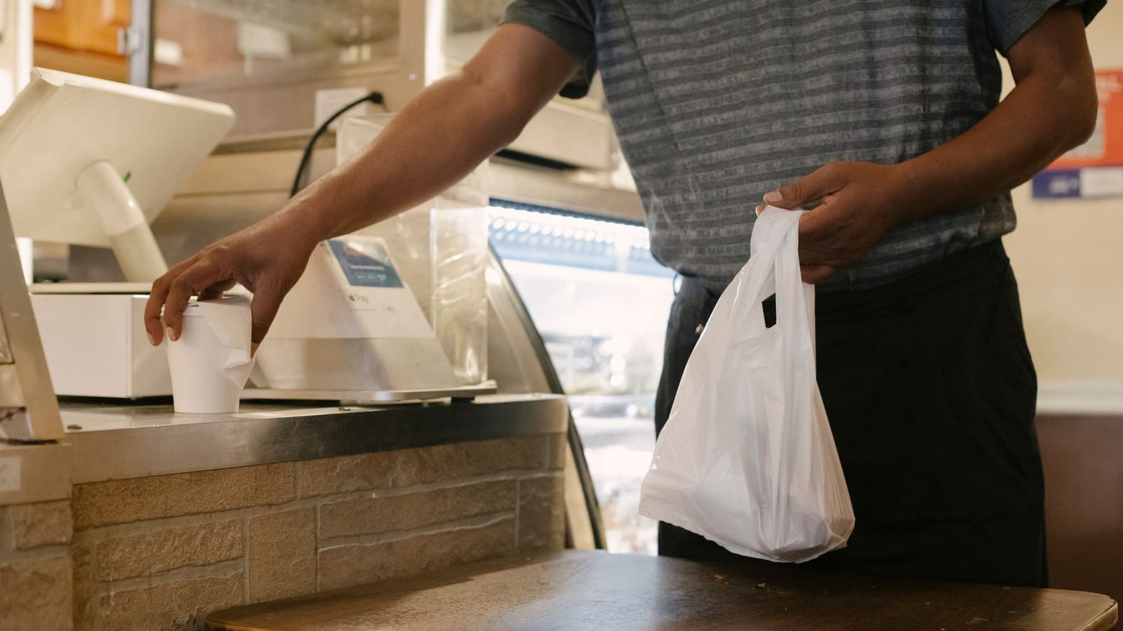 A customer picks up a takeout bag at a cafe counter beside the register and a paper cup of coffee.