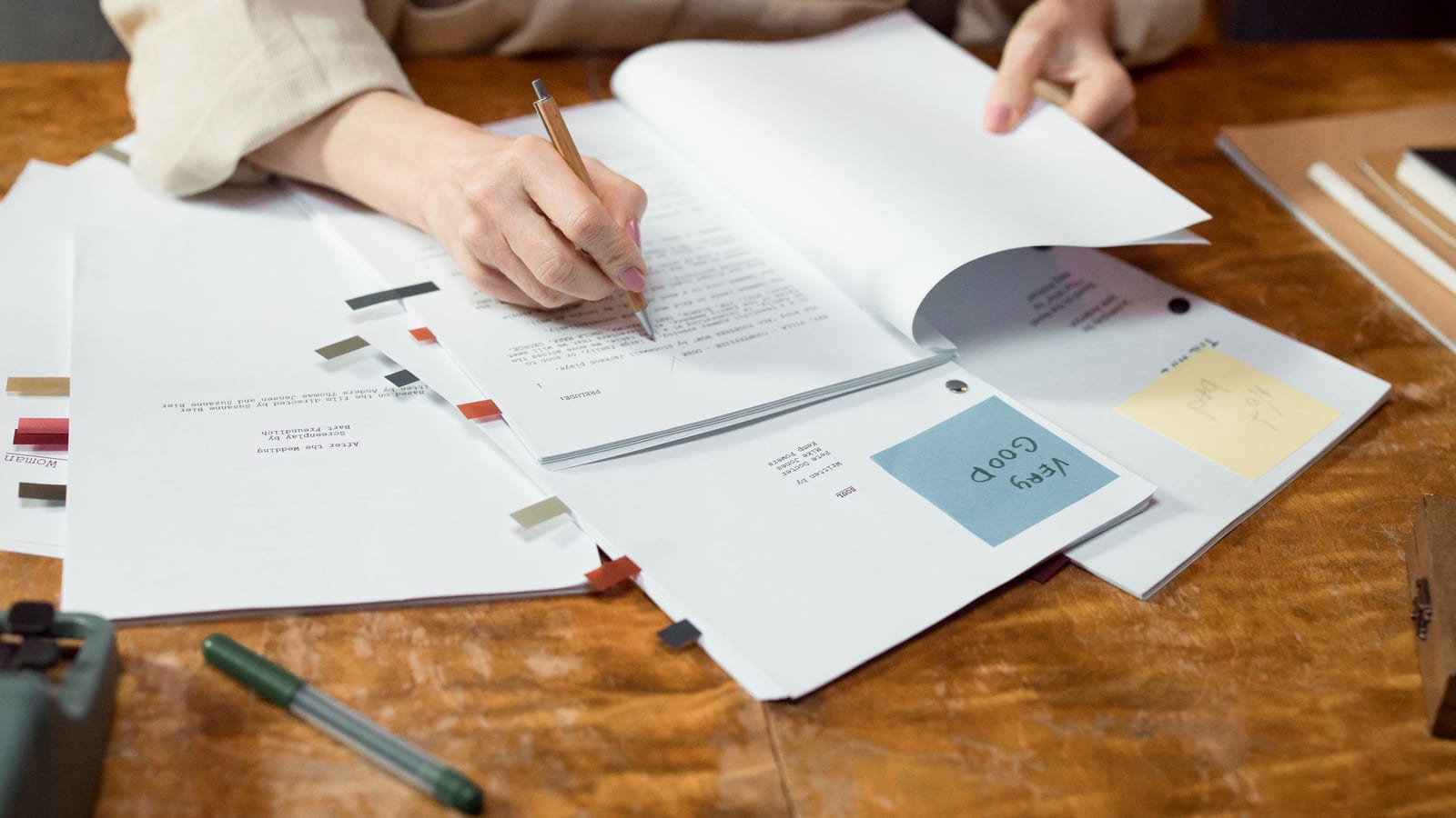 Hands mark up a printed draft with a pen on a wooden desk, more proofs with colored tabs and two handwritten feedback notes spread beside it.