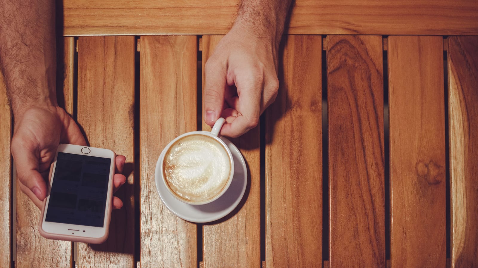 Seen from above, one hand holds a smartphone and the other a cappuccino on a slatted wooden table.