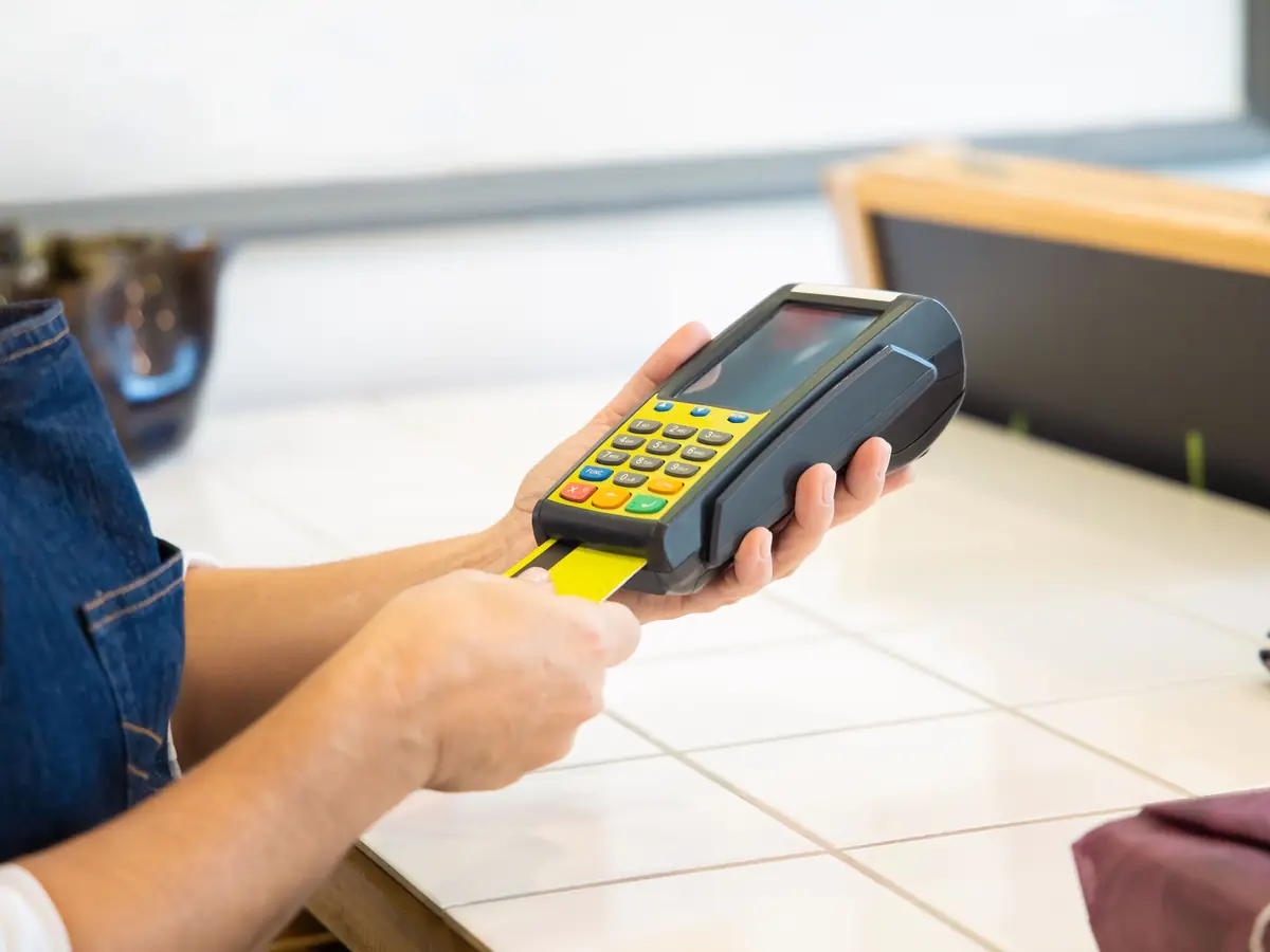 A staff member in a denim apron holds a card reader at a counter while a card goes in.