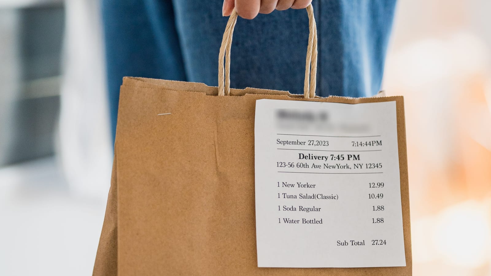 A hand holds a paper takeout bag with a printed delivery order ticket on the front.