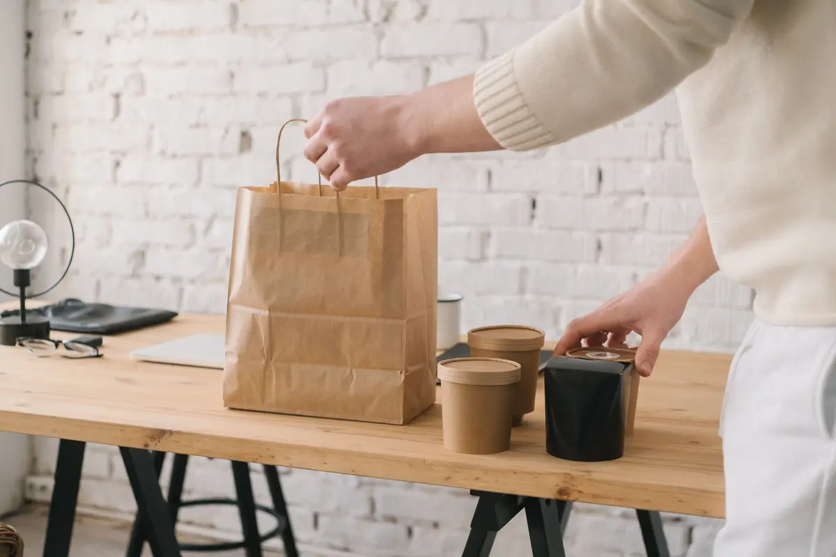 A worker’s hands set lidded takeout cups beside a kraft paper bag on a wooden table.