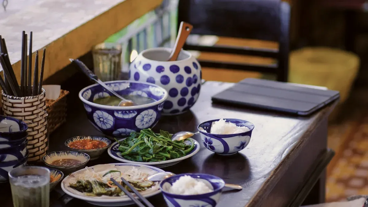 A lunch of soup, greens, rice and dipping sauces in blue-and-white bowls on a dark wooden table, chopsticks in a cup beside them