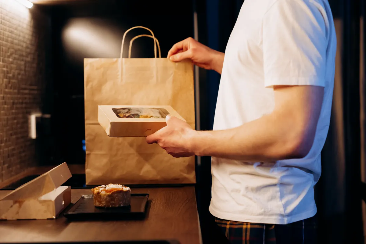 A worker in a white T-shirt packs a box into a brown paper takeout bag at a restaurant counter.