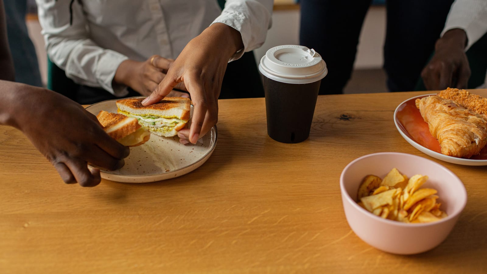 Coworkers in office shirts reach for a catered lunch of sandwiches, crisps and coffee on a wooden table.