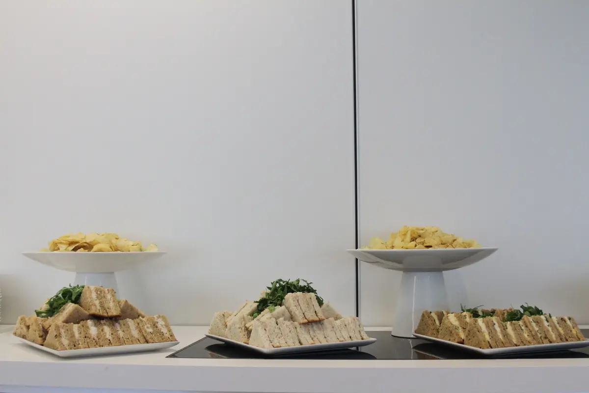 Plates of crustless sandwiches and bowls of crisps set out in a row on a white counter against a plain wall.