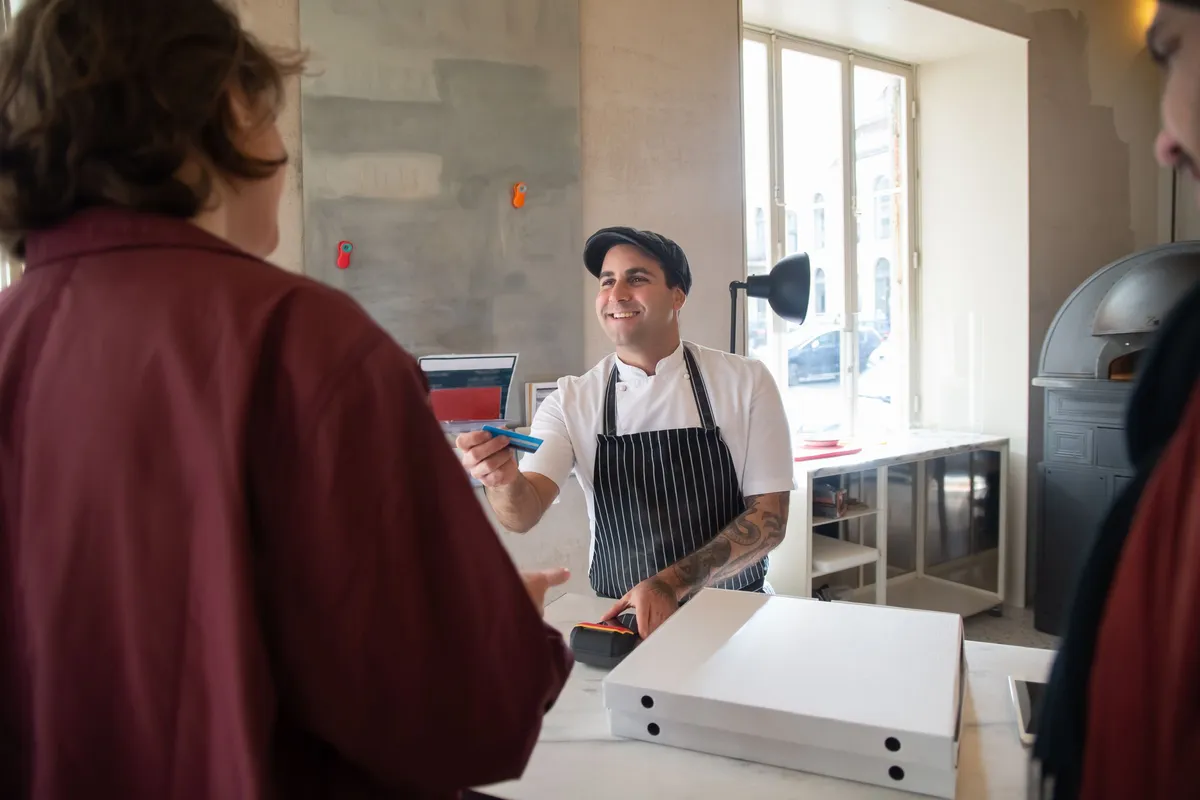 A cook in a striped apron smiles across a pizzeria counter at a customer, a boxed order ready beside him.
