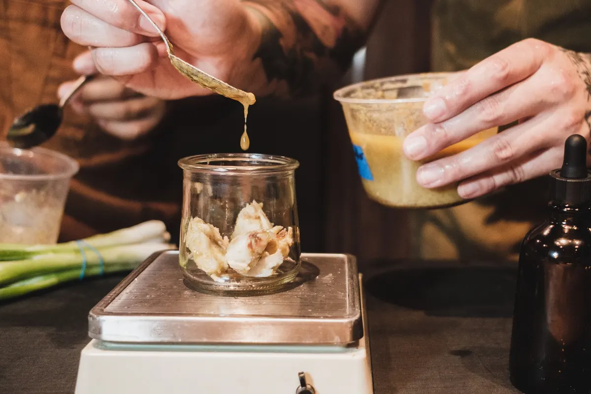 Hands spoon a sauce into a glass jar of ingredients resting on a small kitchen scale.