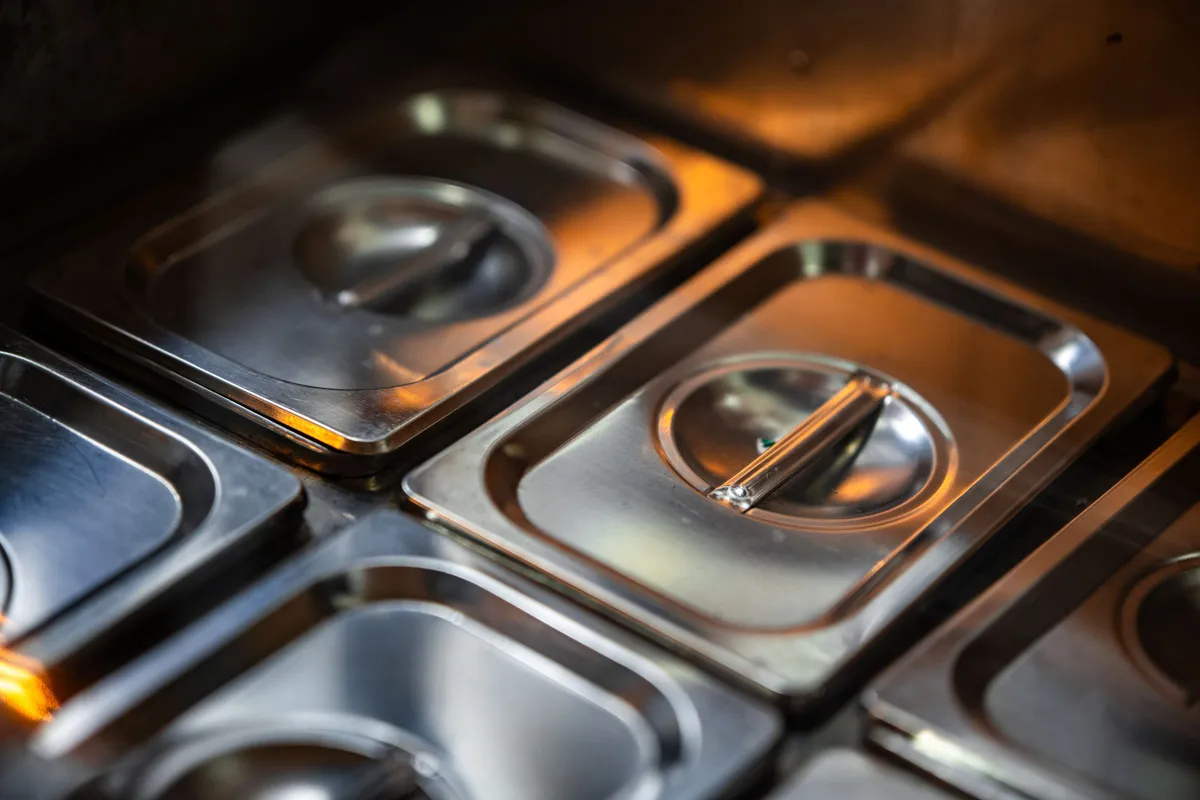 Lidded steel food pans sit in a kitchen line under warm light, ready for a menu that is not yet served.