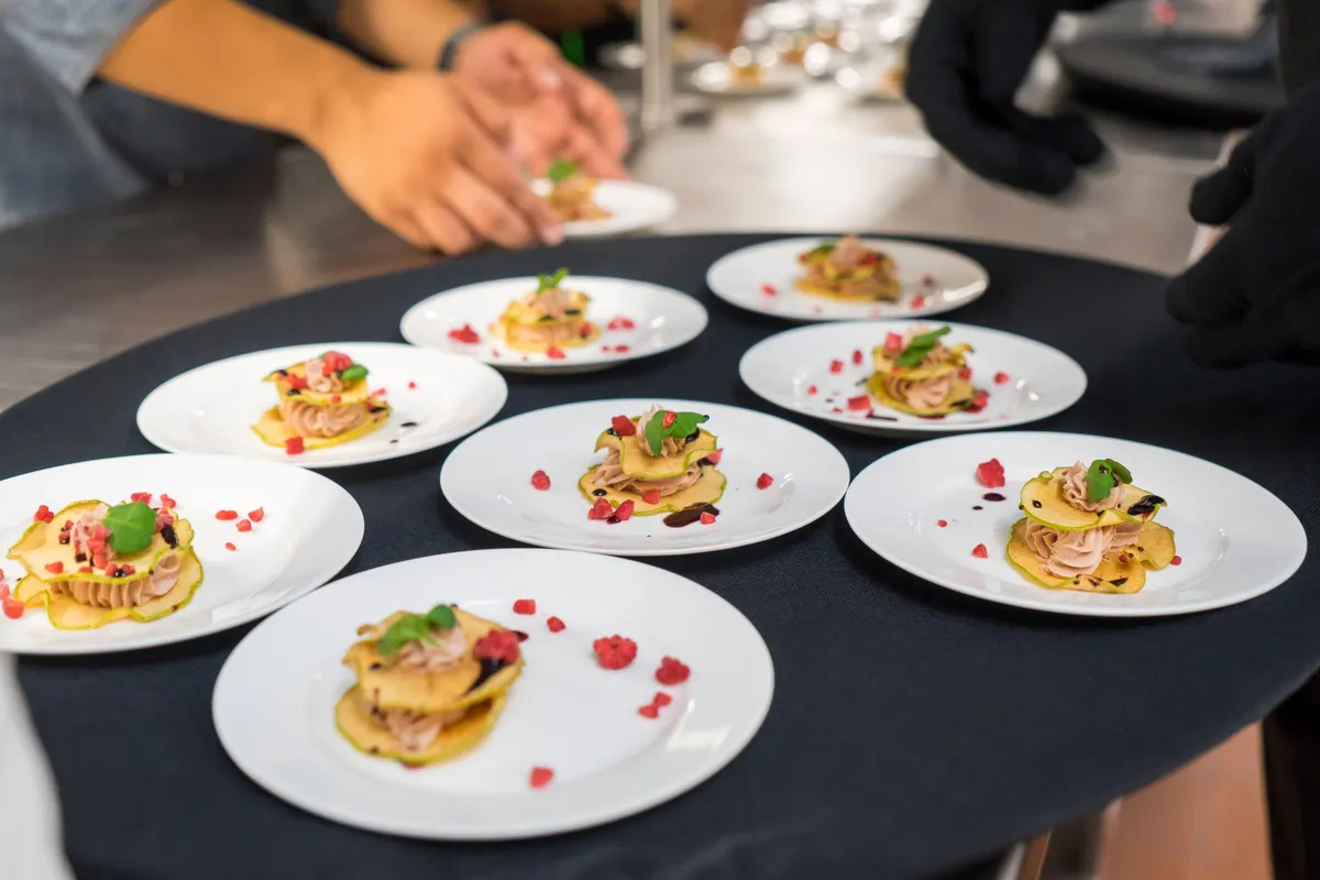A cook sets the last of eight plated tasting dishes on a dark table, the kind of spread a soft opening serves.