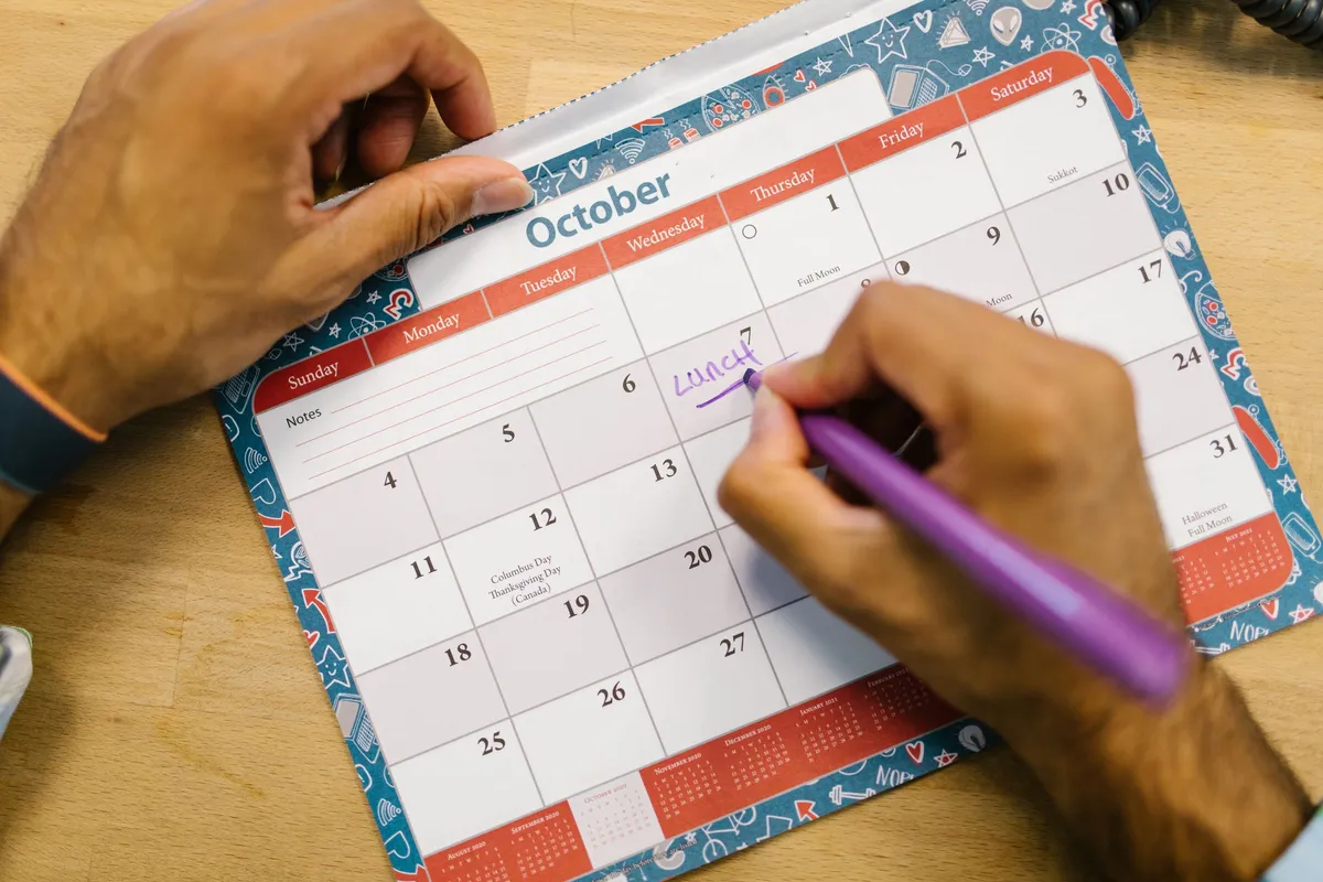 Two hands write a booking into one day of an October desk calendar with a purple marker.