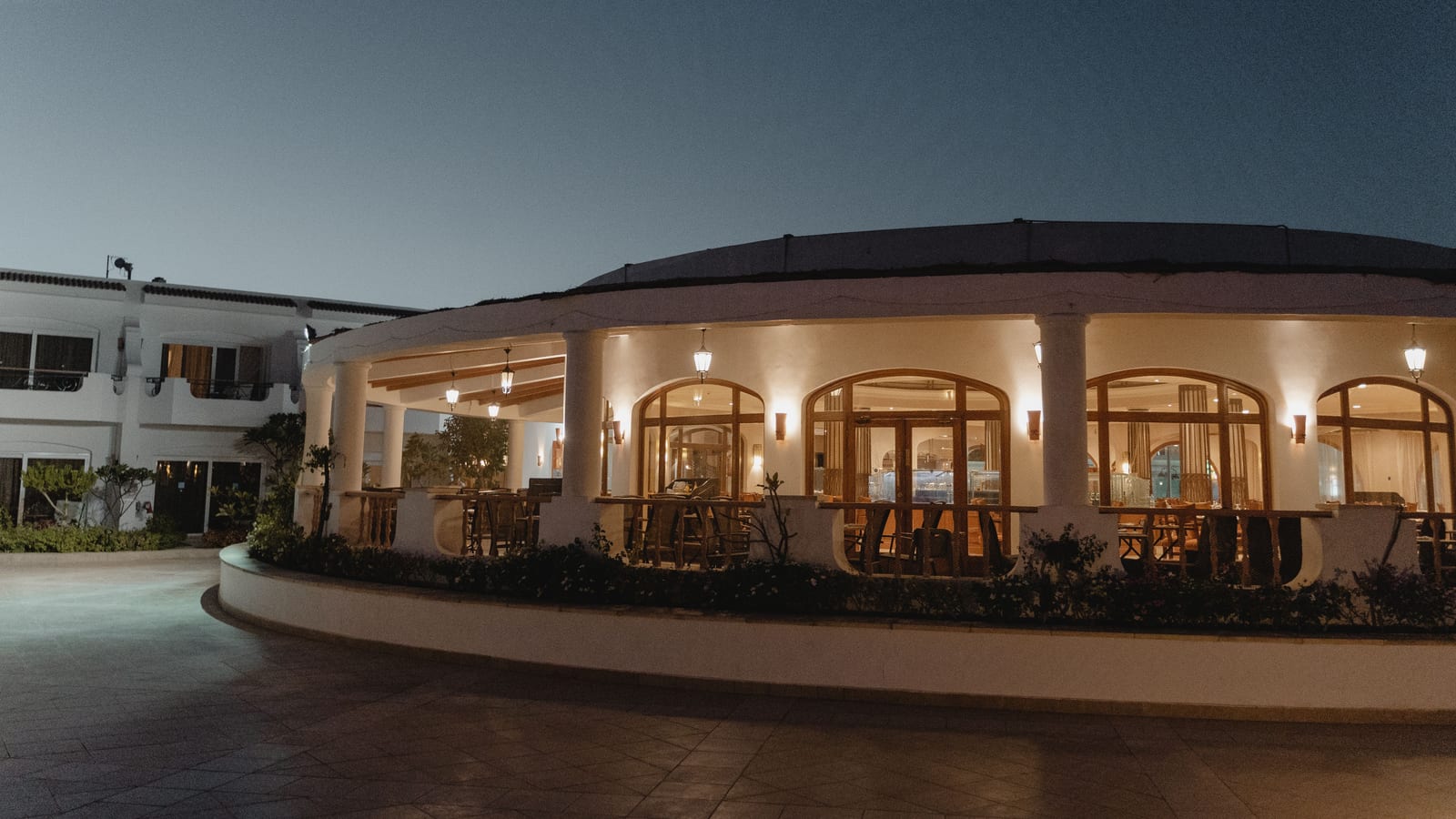 A restaurant location glows at dusk, its arched windows lit over an empty dining room and an empty paved forecourt.