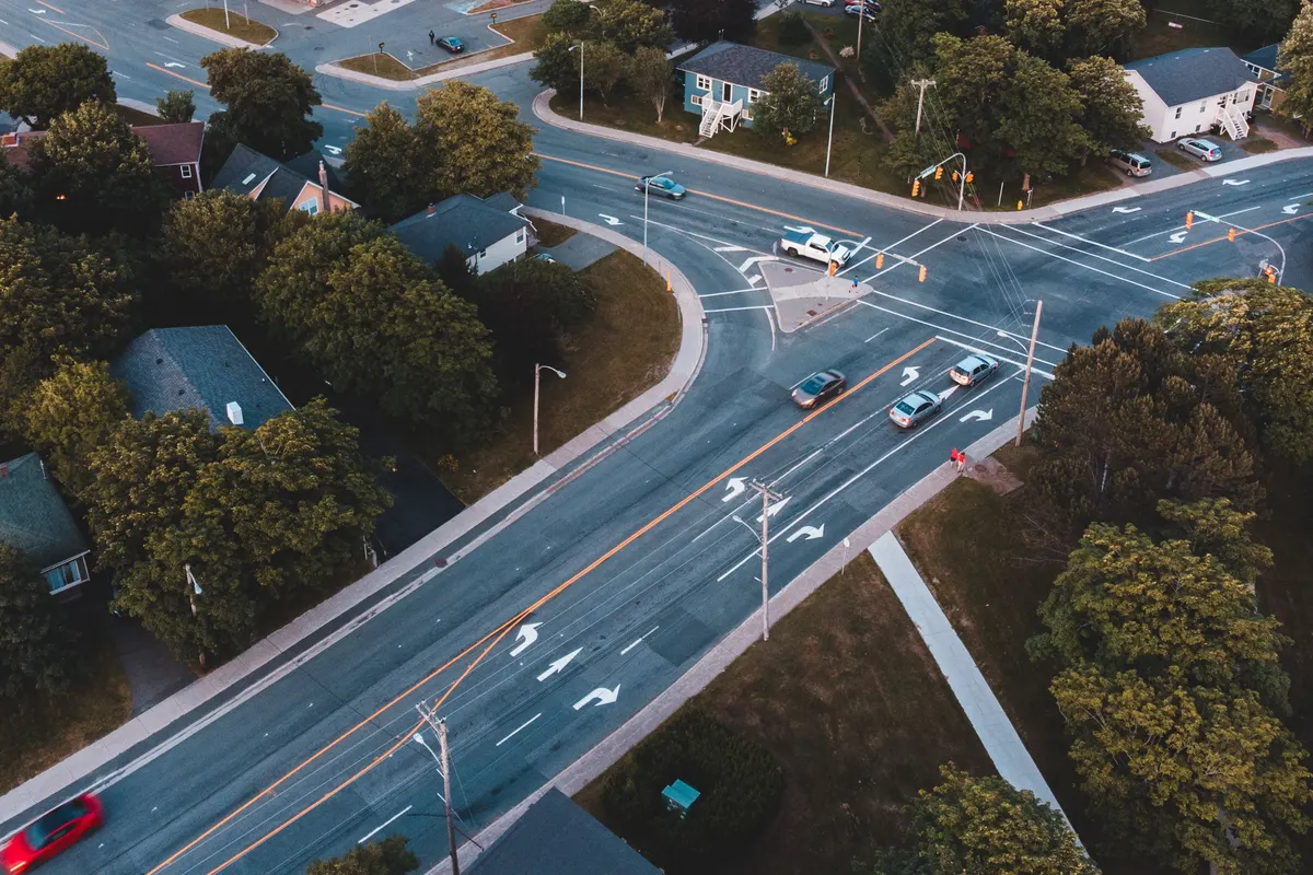An aerial view of a suburban crossroads, where a main road with turn lanes runs past houses and trees.