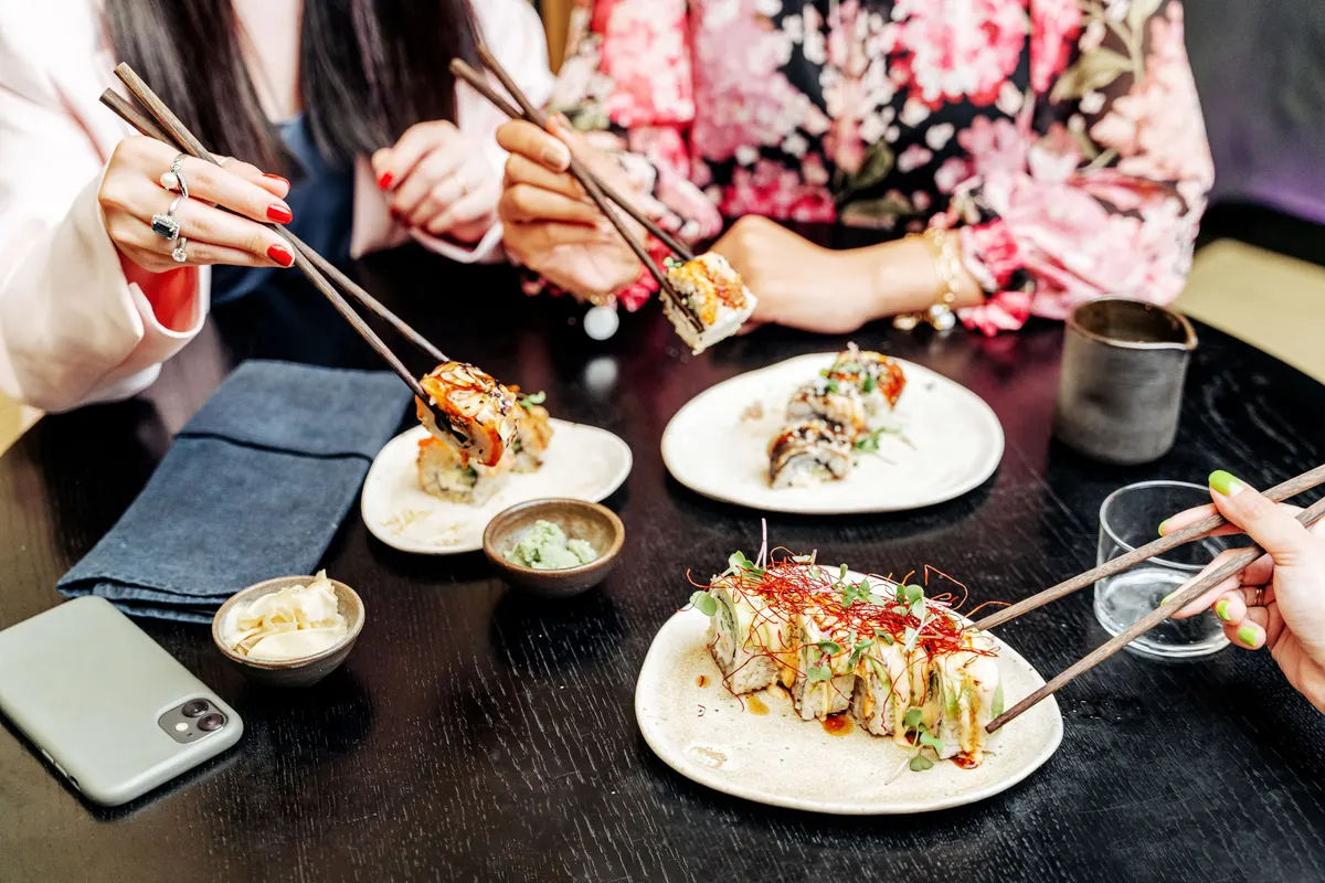 Three friends reach across a restaurant table with chopsticks to share plates of sushi rolls.