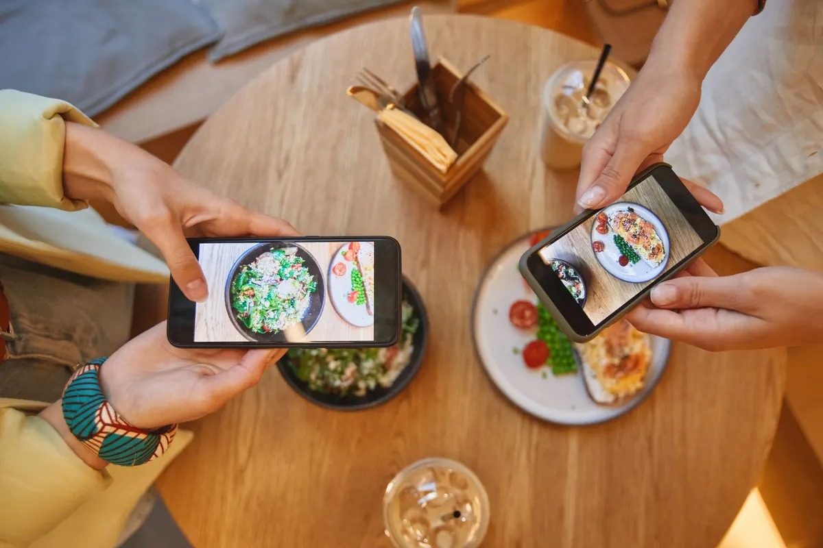 Two pairs of hands hold phones over a cafe table and photograph a salad bowl and a plate of toast.